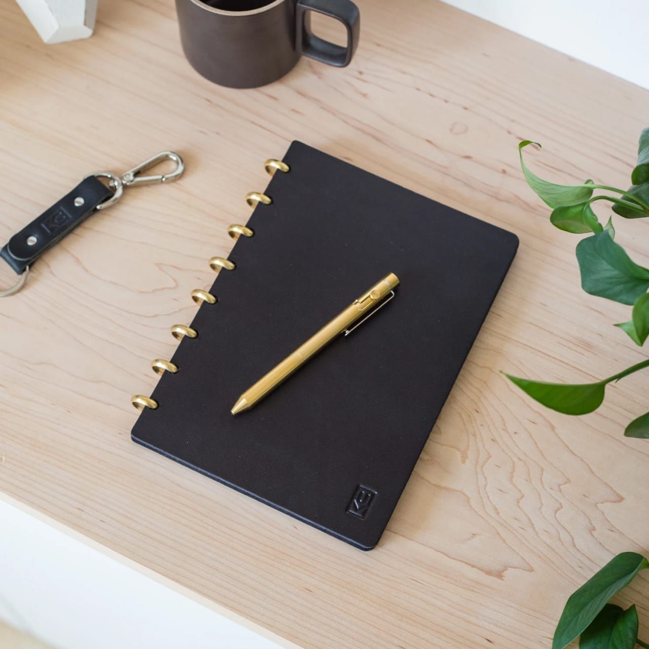 Wood table with porcelain mug, leather journal, brass pen, leather key ring, and a houseplant.